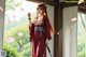 A woman in traditional attire holds a sword, standing near a window with cherry blossom petals.